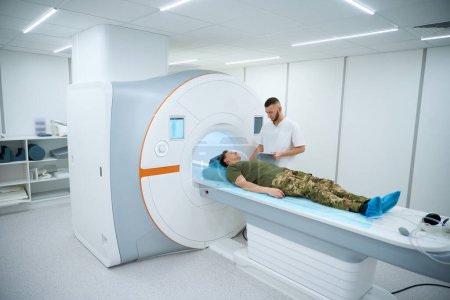 Photo for Radiographer placing shoulder coil over arm of serviceperson positioned on magnetic resonance imaging table - Royalty Free Image