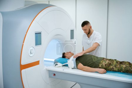 Técnico MR colocando bobina sobre el brazo superior del paciente militar posicionado sobre la mesa de resonancia magnética