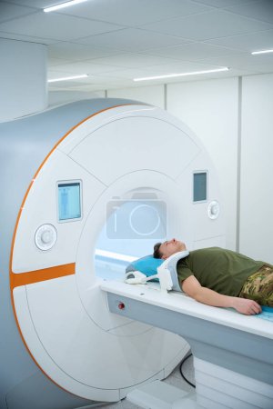 Photo for Military man with coil placed over upper arm lying on back on MRI table in scanning room - Royalty Free Image