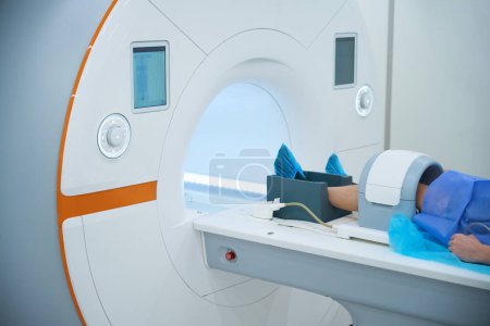 Ausgeschnittenes Foto einer erwachsenen Person mit Spule, die um das Knie gelegt wird, liegend auf einem Magnetresonanztomographen-Tisch