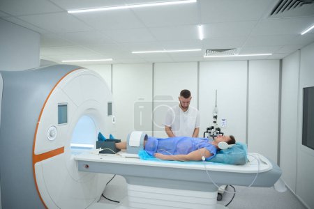 Mann mit geräuschunterdrückenden Kopfhörern und Spule um das Knie liegend auf Magnetresonanztomographen in Anwesenheit eines Röntgenbildners