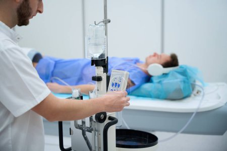 Mann mit Kopfhörer und Spule um das Knie liegend auf Magnetresonanztomographie-Tisch, während Röntgenbild Einstellungen am Kontrastinjektor vornimmt