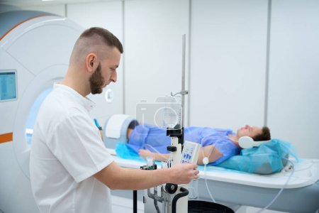 Patient mit Kopfhörer und Spule um das Knie auf Magnetresonanztomographie-Tisch positioniert, während Technologe die Einstellungen am Kontrastinjektor anpasst