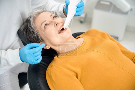 Close-up dentist scanning teeth of mature woman client with intraoral scanner, looking for broken feelings and teeth with decay, checking gums for inflammation