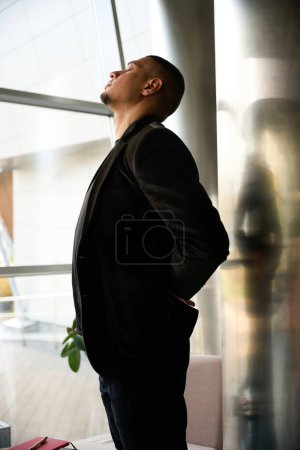 Photo for Side view picture of calm young man standing near window while raising head, closing eyes and folding hands behind back - Royalty Free Image