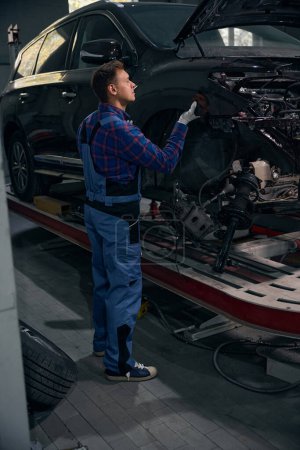 Foto de Hombre en ropa protectora de pie cerca de la máquina e instalando el elemento del cuerpo en el centro de servicio del vehículo - Imagen libre de derechos