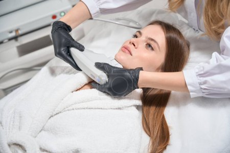 Cropped female beautician doing laser depilation with laser hair removal machine of face of adult caucasian woman in blurred beauty salon. Concept of face skin care