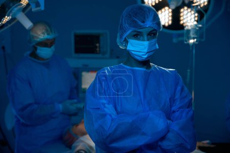 Téléchargez les photos : Médecin confiante en uniforme chirurgical debout dans la salle d'opération moderne sombre - en image libre de droit