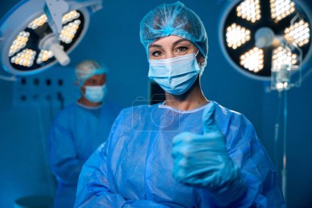 Téléchargez les photos : Joyeux médecin féminin en uniforme chirurgical montrant pouce levé comme geste debout dans la salle d'opération moderne - en image libre de droit