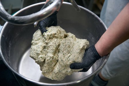 Kitchen worker takes out dough with chopped herbs from a dough kneading machine, the man works in protective gloves