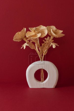 Eine hölzerne Vase mit einem Blumenstrauß aus Stroh auf rotem Hintergrund. Strohflechten