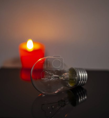 Photo for Burning candle near a switched off light bulb in complete darkness. Blackout, electricity off, energy crisis or power outage, concept image. - Royalty Free Image