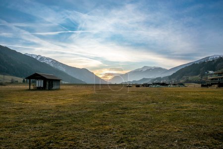 Photo for Sunset behind the snowy mountains in the Austrian Alps. A sunlit meadow in a valley on the edge of a village in Austria. - Royalty Free Image