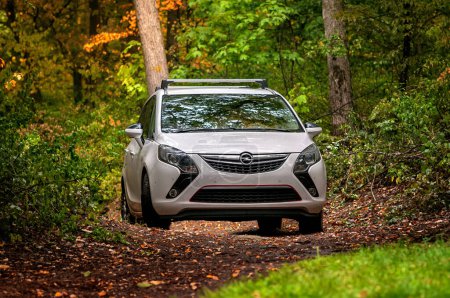 Téléchargez les photos : Voiture familiale blanche MPV Opel Zafira en automne dans la forêt au milieu des arbres. Automne tombant feuilles sur le sol, feuilles vertes et colorées sur les arbres. - en image libre de droit