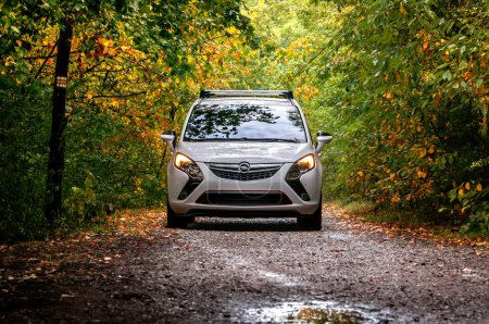 Téléchargez les photos : Voiture familiale blanche MPV Opel Zafira en automne dans la forêt au milieu des arbres. Automne tombant feuilles sur le sol, feuilles vertes et colorées sur les arbres. - en image libre de droit