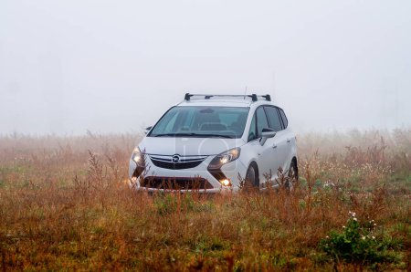 Téléchargez les photos : Brno, République tchèque - 6 novembre 2023 : Une Opel Zafira blanche se dresse dans un champ d'herbe beige à l'automne, avec ses lumières allumées. Il y a du brouillard en arrière-plan. - en image libre de droit