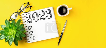 Foto de Resoluciones de año nuevo 2023 en el escritorio. Lista de resoluciones 2023 con cuaderno, taza de café sobre mesa amarilla. Objetivos, resoluciones, plan, acción, concepto de lista de verificación. Plantilla de año nuevo 2023, copia spac - Imagen libre de derechos