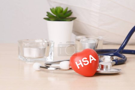 la palabra hsa está escrita en el juguete rojo en forma de corazón sobre una mesa de madera cerca de un estetoscopio en el fondo