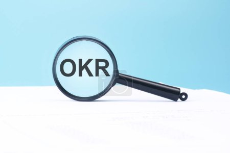 OKR-Text auf Lupe auf blauem und weißem Hintergrund, Geschäftskonzept