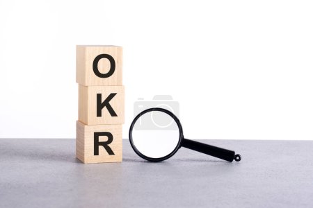 OKR - Ziele und Schlüsselergebnisse - Text Holzwürfelblöcke und Lupe auf grauem Tisch, Geschäftskonzept