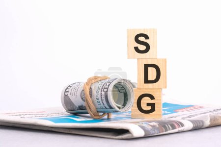 Holzwürfelblock mit dem Text der SDG auf dem Hintergrund einer Zeitung und Banknoten. Finanz-, Management-, Wirtschafts-, Geschäftskonzept