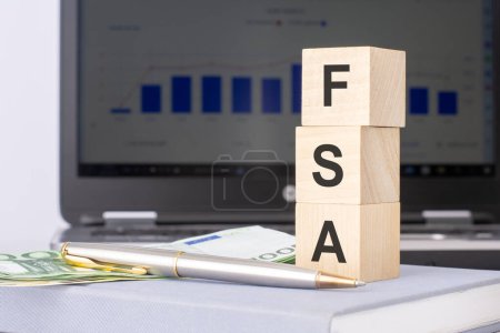 Nahaufnahme von Holzklötzen mit dem Akronym FSA und Banknoten auf dem Hintergrund eines Laptops, Geschäftskonzept