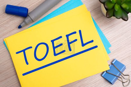 toefl - Abkürzung für Test Of English As A Foreign Language. Text auf gelbem Papier auf hellem Holzgrund mit Papeterie