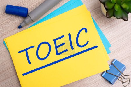 toeic - short for Test Of English For International Communication. text on yellow paper on light wooden background with stationery
