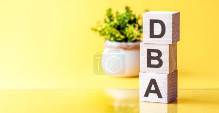 Buchstaben des Alphabets von dba auf Holzklötzen, grüne Pflanze auf gelbem Hintergrund. dba - Abkürzung für Data Base Administrator