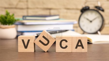 Photo for VUCA is written on the wooden cubes. in the background there is an clock near a potted plant and a stack of books - Royalty Free Image