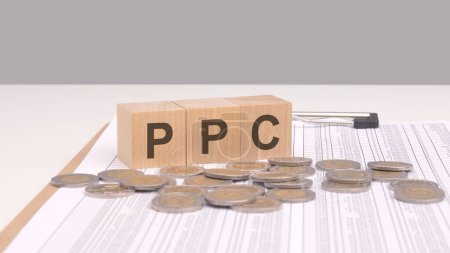 Texto PPC en bloques de madera sobre una mesa de madera con monedas de oro. PPC significa Pay-Per-Click, un modelo de marketing digital donde los anunciantes pagan una tarifa cada vez que se hace clic en su anuncio.