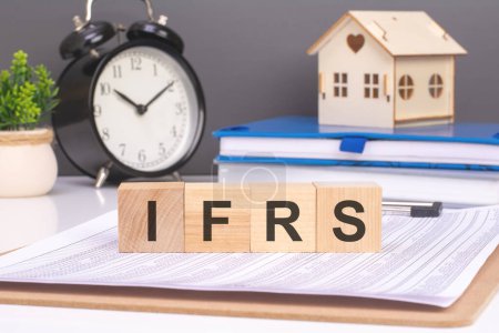 arrangement wooden blocks spelling IRFS along side a small wooden house and alarm clock on a gray background. represent the adoption of these global financial reporting standards