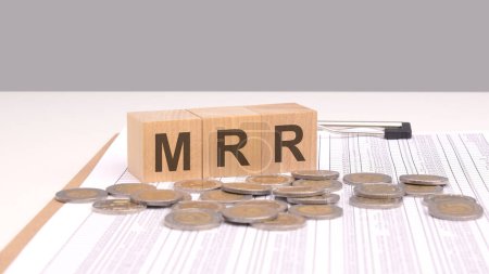 Texto MRR en bloques de madera sobre una mesa blanca con monedas. MRR significa Monthly Recurring Revenue, una métrica clave en los modelos de negocio basados en suscripción, que representa el ingreso predecible y estable..