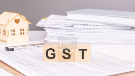 Holzwürfel mit Text GST auf einem Dokument mit Tabellen. Hintergrund: eine hölzerne Hausfigur und ein Aktenstapel. Konzept: Waren- und Dienstleistungssteuer