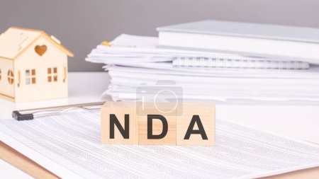 wooden cubes with text nda on a document with tables. Background: a wooden house figure and document stack. Concept: Non-Disclosure Agreement