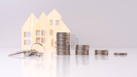 Der Schlüssel liegt neben einem Stapel Münzen vor den kleinen Modellen auf grauem Hintergrund. Musterhaus aus Holz mit Schlüssel und Münzen - Immobilieninvestitionskonzept