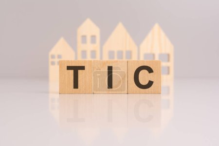 Holzklötze, die den Text "TIC" auf grauem Hintergrund mit einem Miniatur-Holzmodellhaus bilden. Spiegelung auf der Tischplatte. "TIC" steht für "Mietrecht in Common"