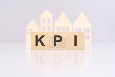 wooden blocks forming the text 'KPI' on a gray background with a miniature wooden model house. reflection on the tabletop. 'KPI' stands for 'Key Performance Indicators'