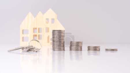 Der Schlüssel liegt neben einem Stapel Münzen vor den kleinen Modellen auf grauem Hintergrund. Musterhaus aus Holz mit Schlüssel und Münzen - Immobilieninvestitionskonzept