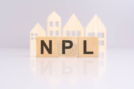 Holzblöcke, die den Text "NPL" auf grauem Hintergrund mit einem Miniatur-Holzmodellhaus bilden. Spiegelung auf der Tischplatte. "NPL" steht für "Non Performing Loans".
