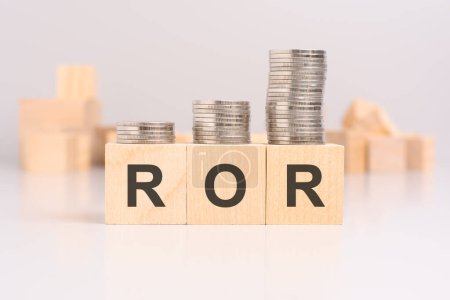 Holzklötze mit dem Wort ROR und gestapelten Münzen auf hellem Hintergrund, im Hintergrund ein verschwommenes Bild vieler zufällig angeordneter Holzwürfel