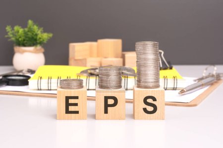 closeup of EPS text written on wooden blocks with stacked coins. office desk with yellow notepad, pen and magnifying glass in the background.