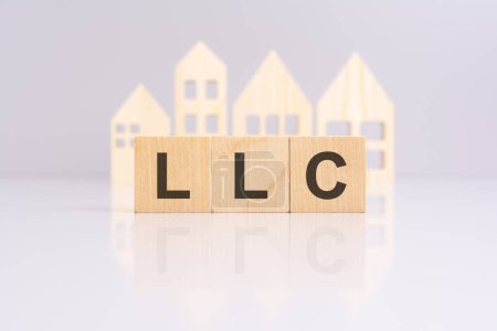 wooden blocks forming the text LLC on a gray background with a miniature wooden model house. reflection on the tabletop. LLC stands for Limited Liability Company