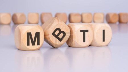 four wooden blocks with the letters MBTI standing at an angle to the viewer on a light gray table and reflecting off the surface. there are many cubes with no inscription in the background