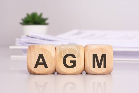 wooden cubes spelling out 'AGM' on the office table, with a white paper document and green flowers in the background, conveys a business concept
