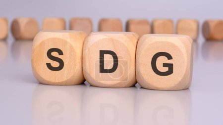 the image depicts three wooden cubes with the letters 'SDG' in focus, reflecting on the table surface. in the background, there is a row of wooden cubes, blurred and out of focus