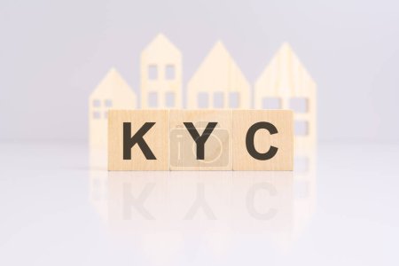 Holzklötze, die den Text "KYC mit einem Miniatur-Holzmodellhaus bilden. Spiegelung auf der Tischplatte. 'KYC' steht für 'Know Your Client'