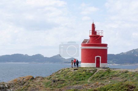 Téléchargez les photos : Les touristes prennent des photos près du phare rouge Punta Robaleira sur le fond de la mer et de l'île avec de hautes montagnes. Voyages en Espagne. Concept de loisirs entre amis. - en image libre de droit