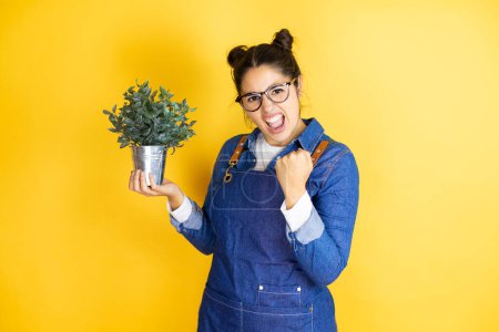 Photo for Young caucasian gardener woman holding a plant isolated on yellow background very happy and excited making winner gesture with raised arms, smiling and screaming for success. - Royalty Free Image