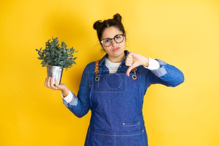 Joven jardinero caucásico mujer sosteniendo una planta aislada sobre fondo amarillo con la cara enojada, signo negativo que muestra aversión con el pulgar hacia abajo